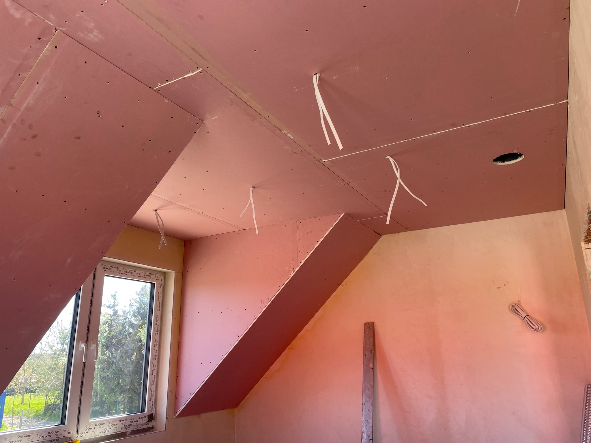 Poddasze w trakcie prac wykończeniowych: zamontowane płyty gipsowo-kartonowe na skosach i suficie, wystające kable elektryczne, widoczne okno z widokiem na zieleń.