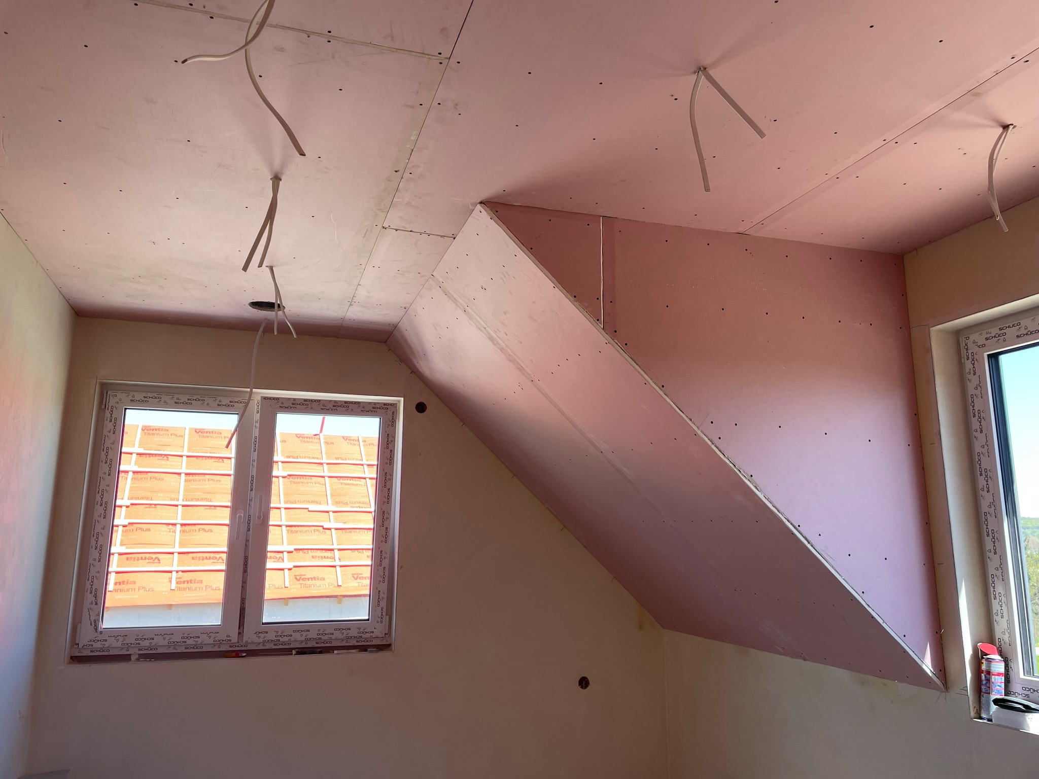 Poddasze w trakcie wykańczania płytami kartonowo-gipsowymi, widoczne wystające kable oświetleniowe i okno dachowe z widokiem na zewnątrz, całość w jasnych barwach.