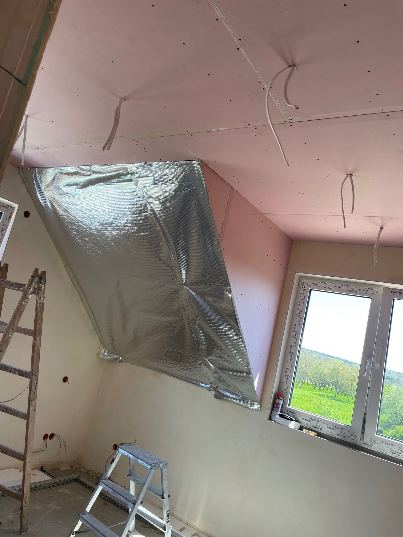 Ocieplenie poddasza folią aluminiową w Mielęcinie, widok na okno z zielonym krajobrazem, drabiny w pomieszczeniu ze ścianami przygotowanymi pod płyty kartonowo-gipsowe.