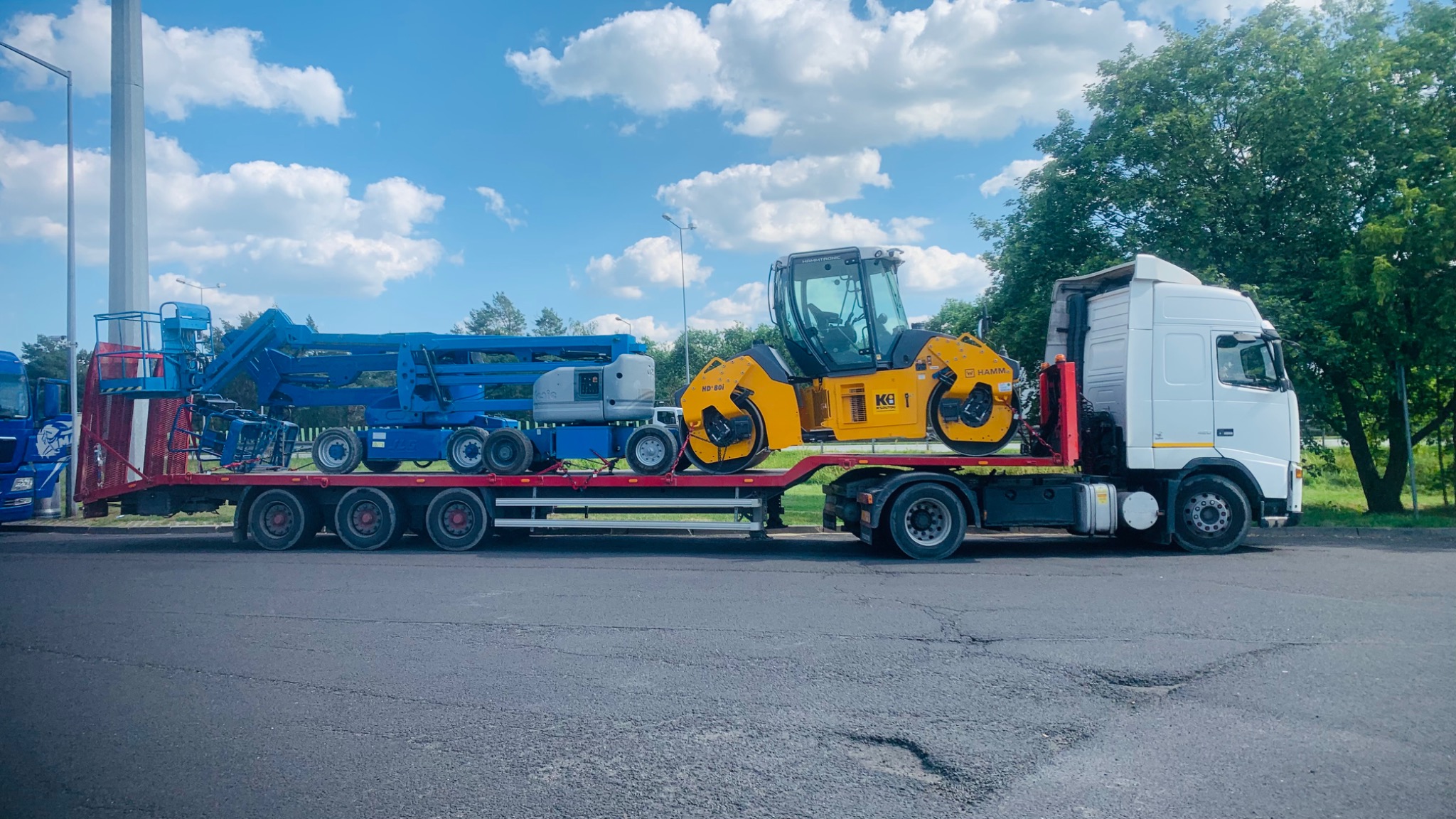 Ciężarówka z naczepą przewożąca niebieski podnośnik koszowy i żółty walec drogowy marki Hamm na tle błękitnego nieba z chmurami.