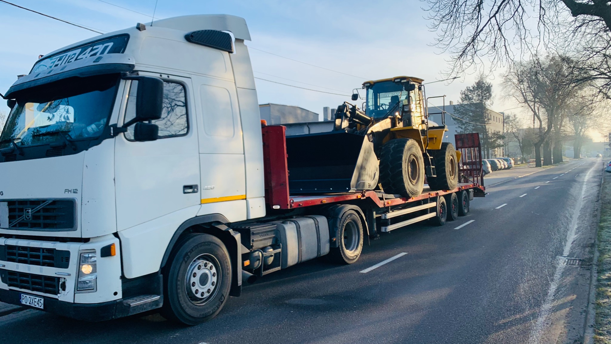 Biała ciężarówka Volvo FH12 z żółtą ładowarką kołową na niskopodwoziowej platformie transportowej, jadąca asfaltową drogą w kierunku światła porannego słońca.