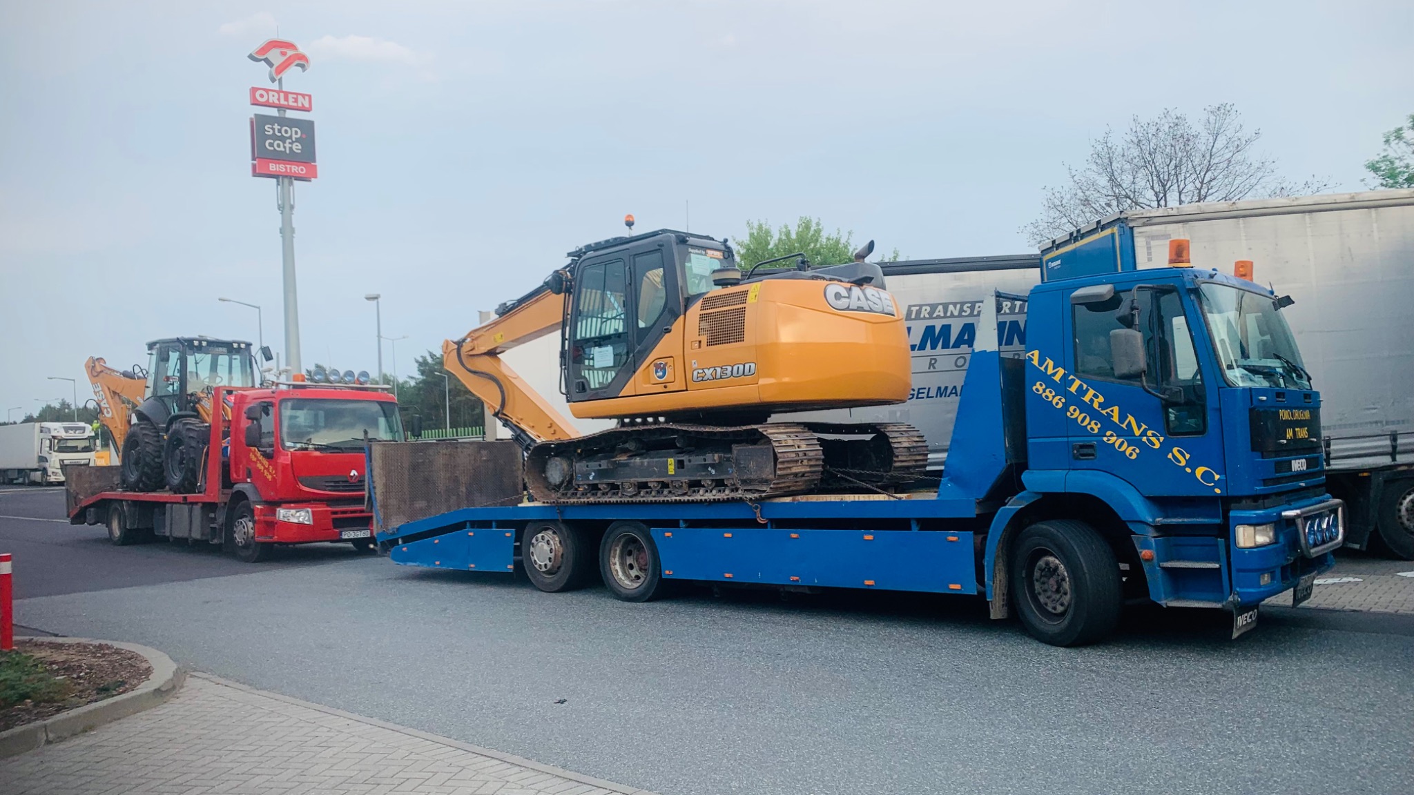 Transport dwóch maszyn budowlanych, koparki CX130D i koparko-ładowarki CASE, na lawetach ciężarowych AM TRANS S.C. na tle stacji Orlen Stop Cafe Bistro.