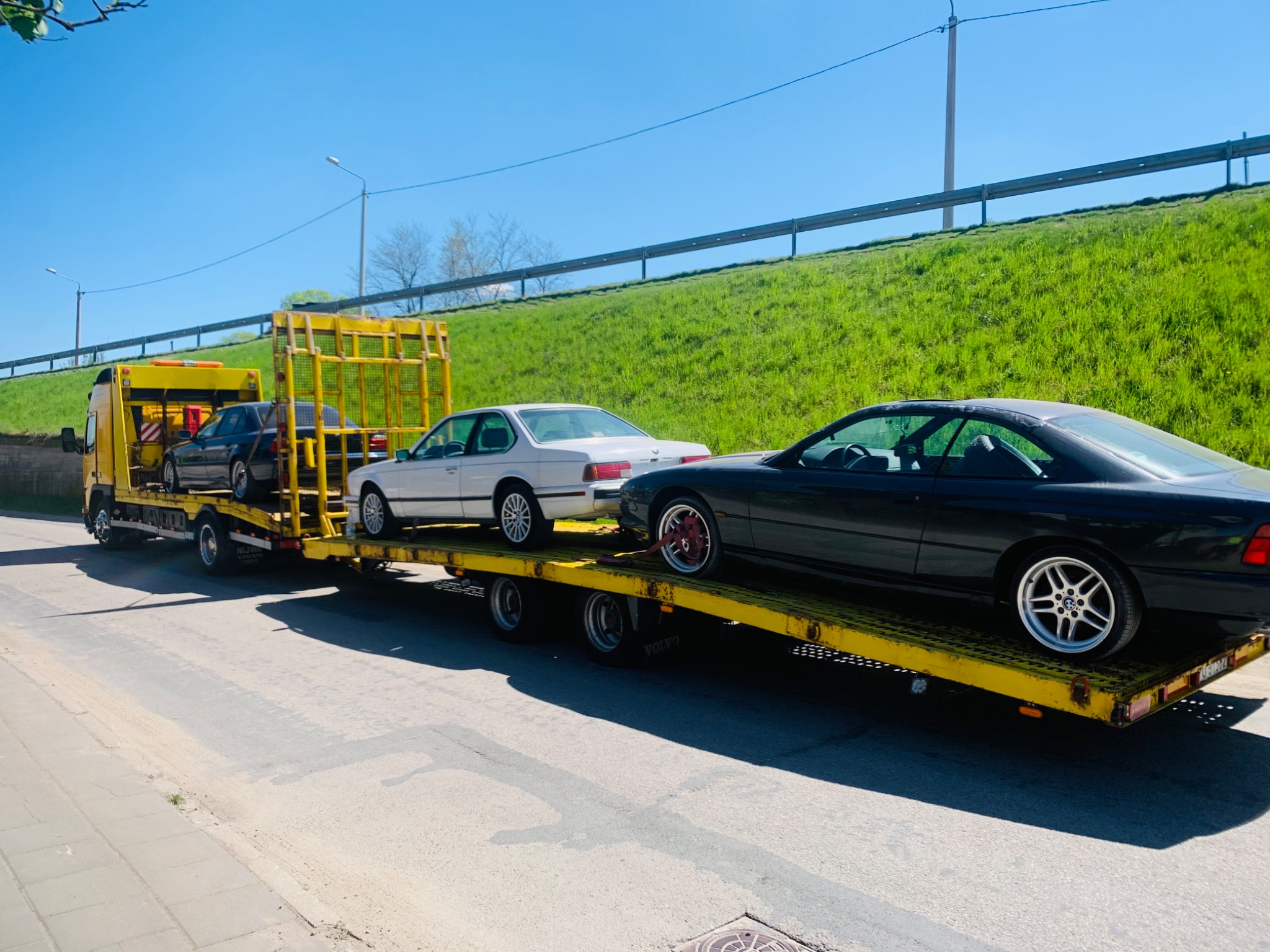 Żółta laweta przewozi trzy samochody BMW serii 6 E24 i E31 w słoneczny dzień. Jeden z samochodów ma czerwone felgi.