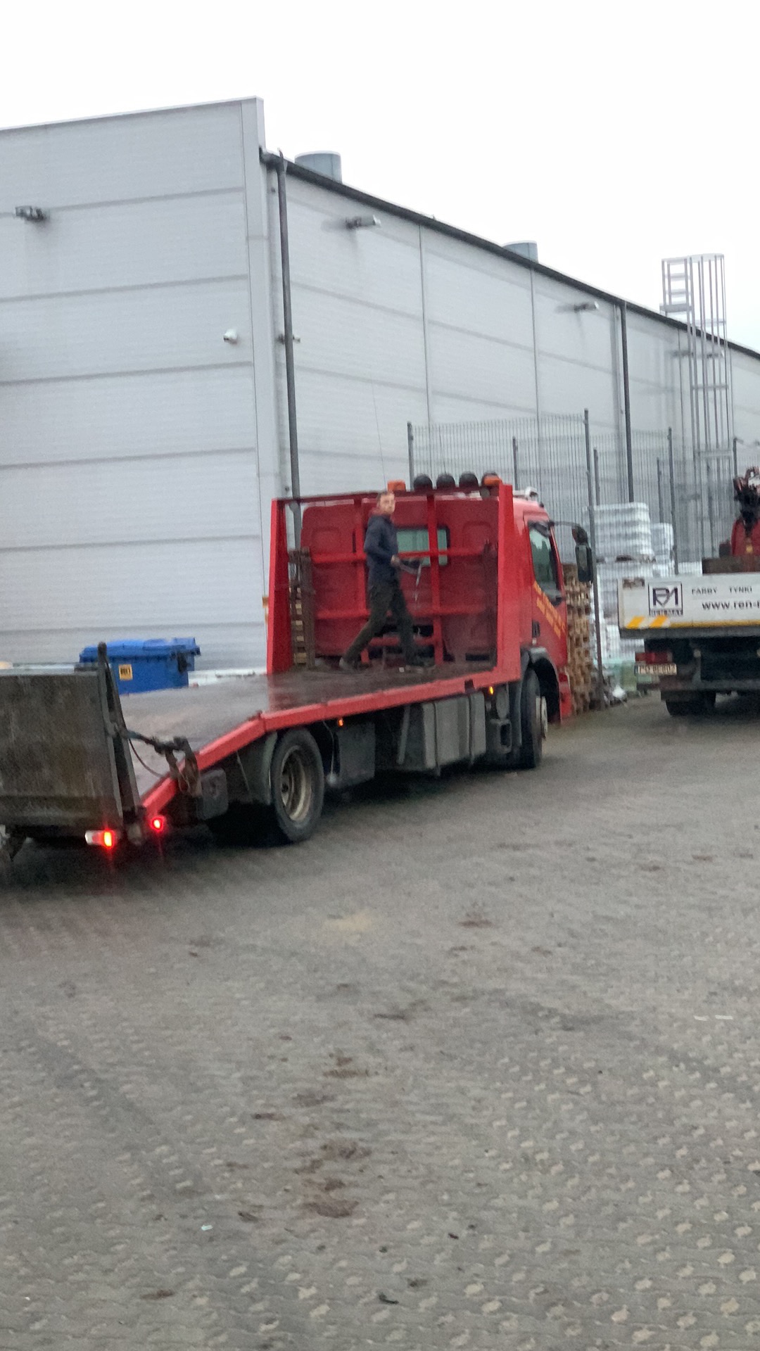 Czerwona laweta z opuszczoną rampą, na której stoi mężczyzna w ciemnym ubraniu, na tle szarego magazynu i innego pojazdu.