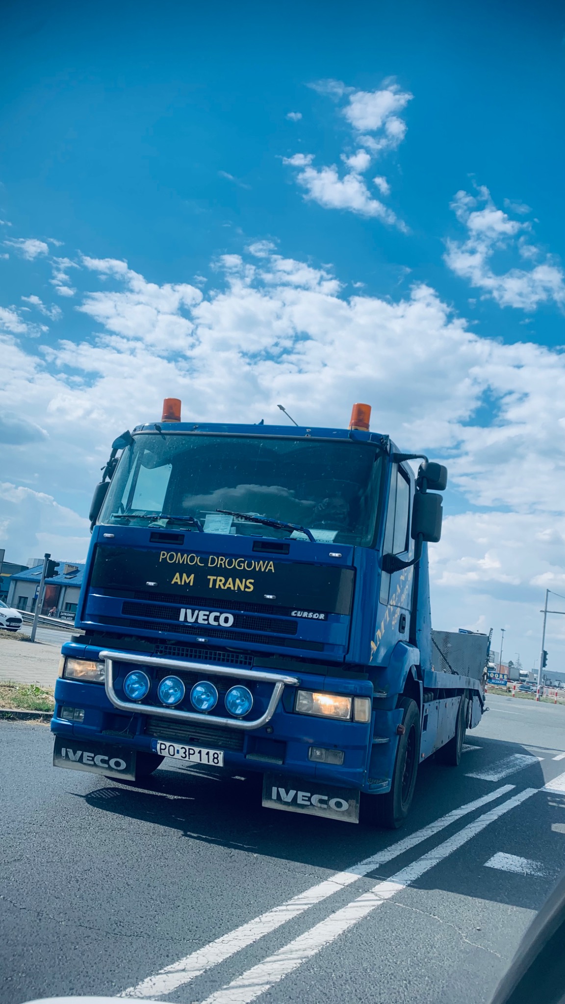 Niebieski samochód pomocy drogowej Iveco AM Trans na drodze, widok z przodu, błękitne niebo z chmurami w tle.