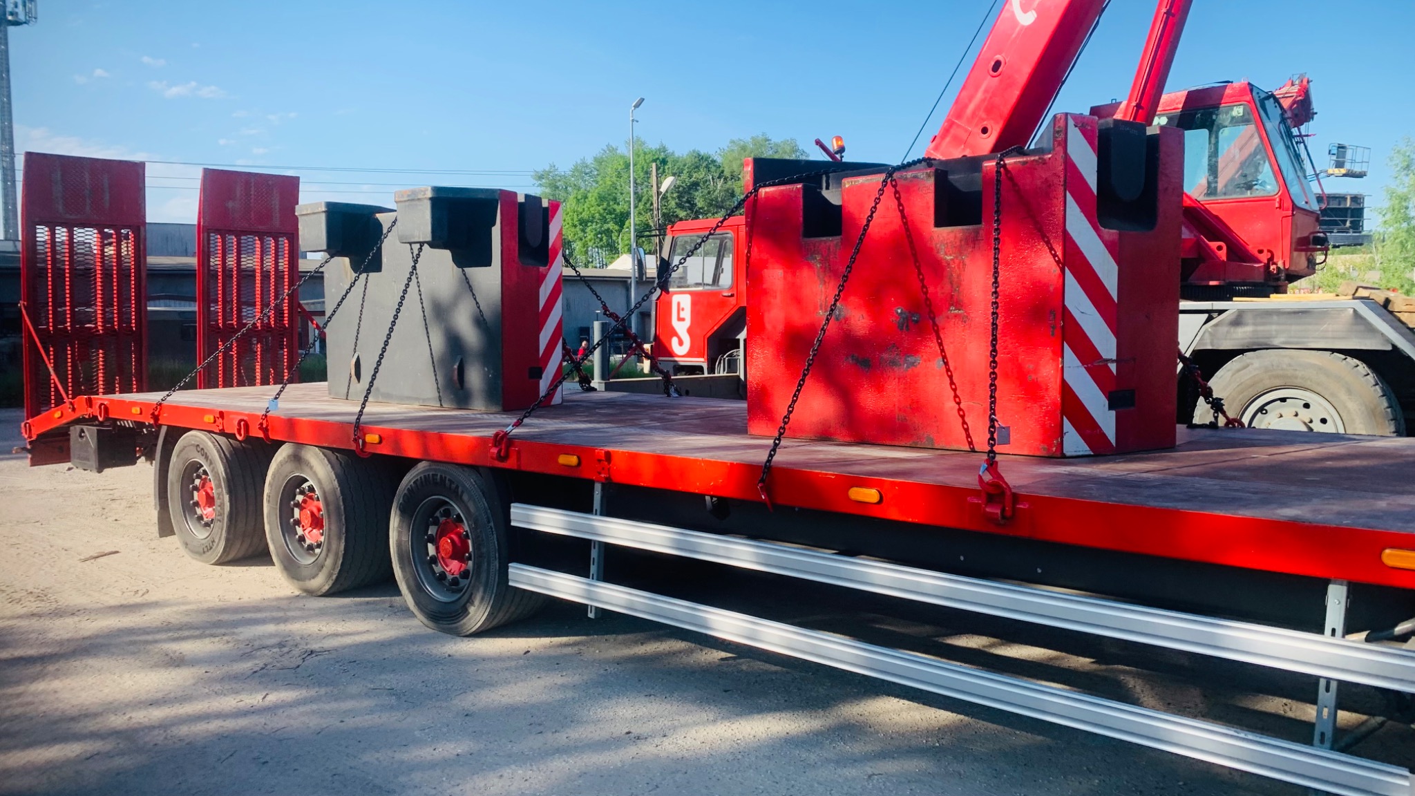 Czerwona naczepa niskopodwoziowa z ładunkiem w postaci ciężkich, szarych bloków zabezpieczonych łańcuchami, z czerwoną kabiną dźwigu w tle.