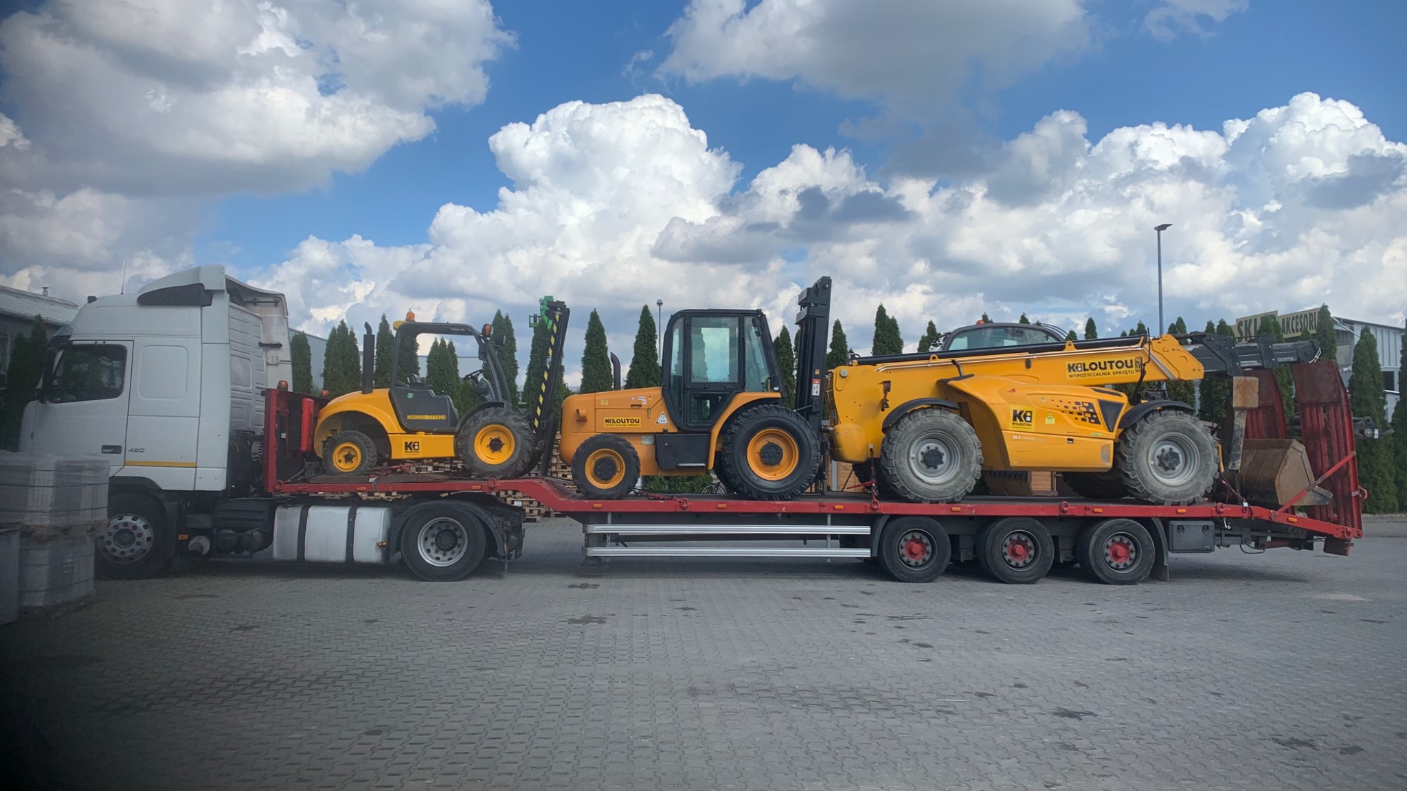 Transport ciężarowy: Biała ciężarówka z naczepą przewozi trzy żółte wózki widłowe marki Kiloutou na tle błękitnego nieba z chmurami.