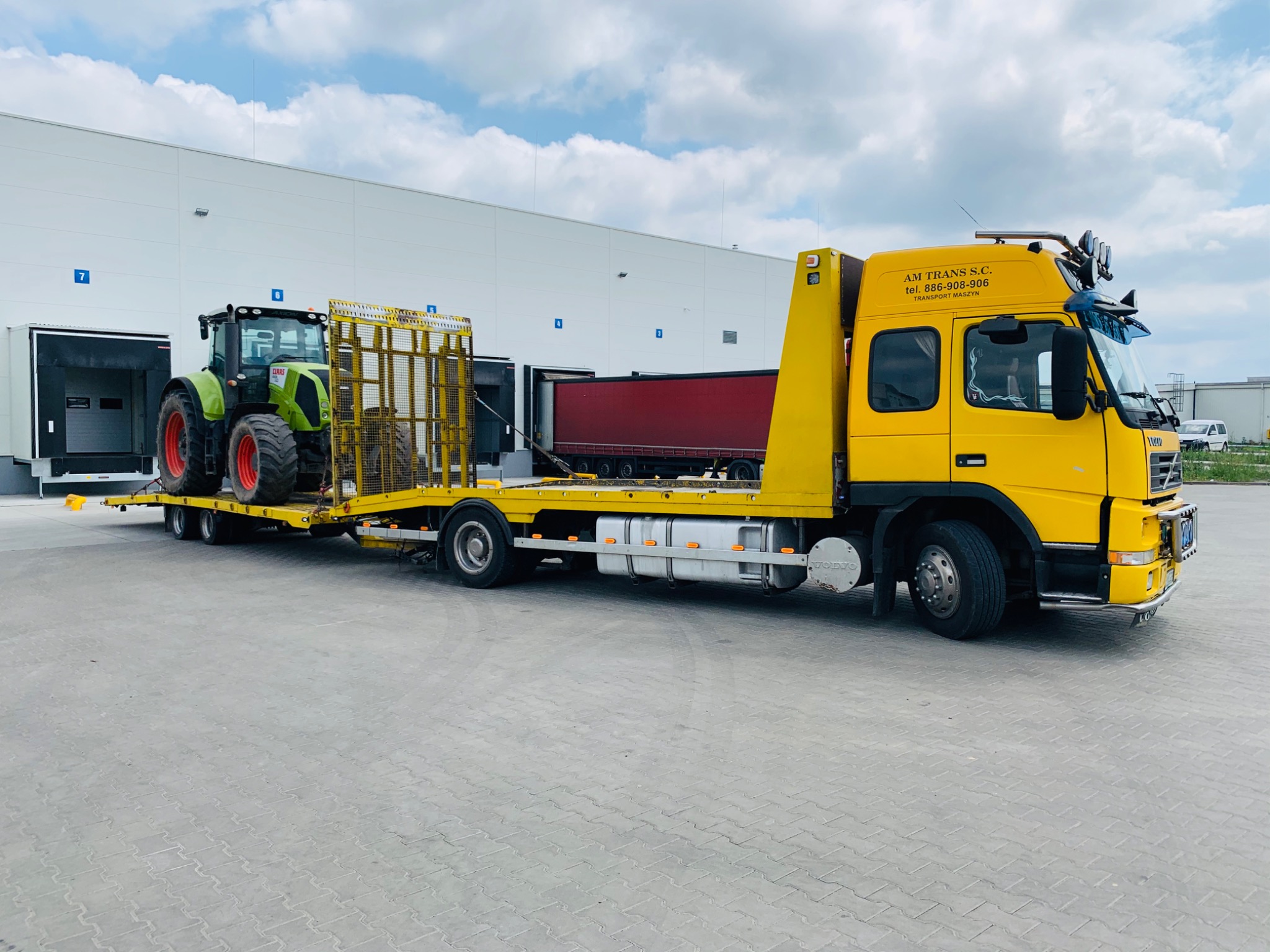 Żółta laweta Volvo z zielonym traktorem ClaaS na platformie, zaparkowana przed magazynem z rampami załadunkowymi.