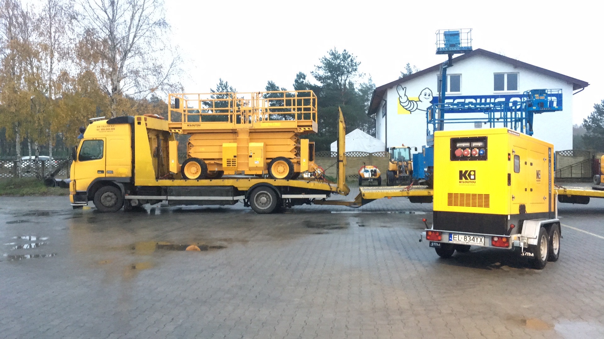 Żółta ciężarówka z platformą transportową przewożąca podnośnik nożycowy i agregat prądotwórczy na przyczepie, na mokrym placu z widocznym budynkiem z reklamą.