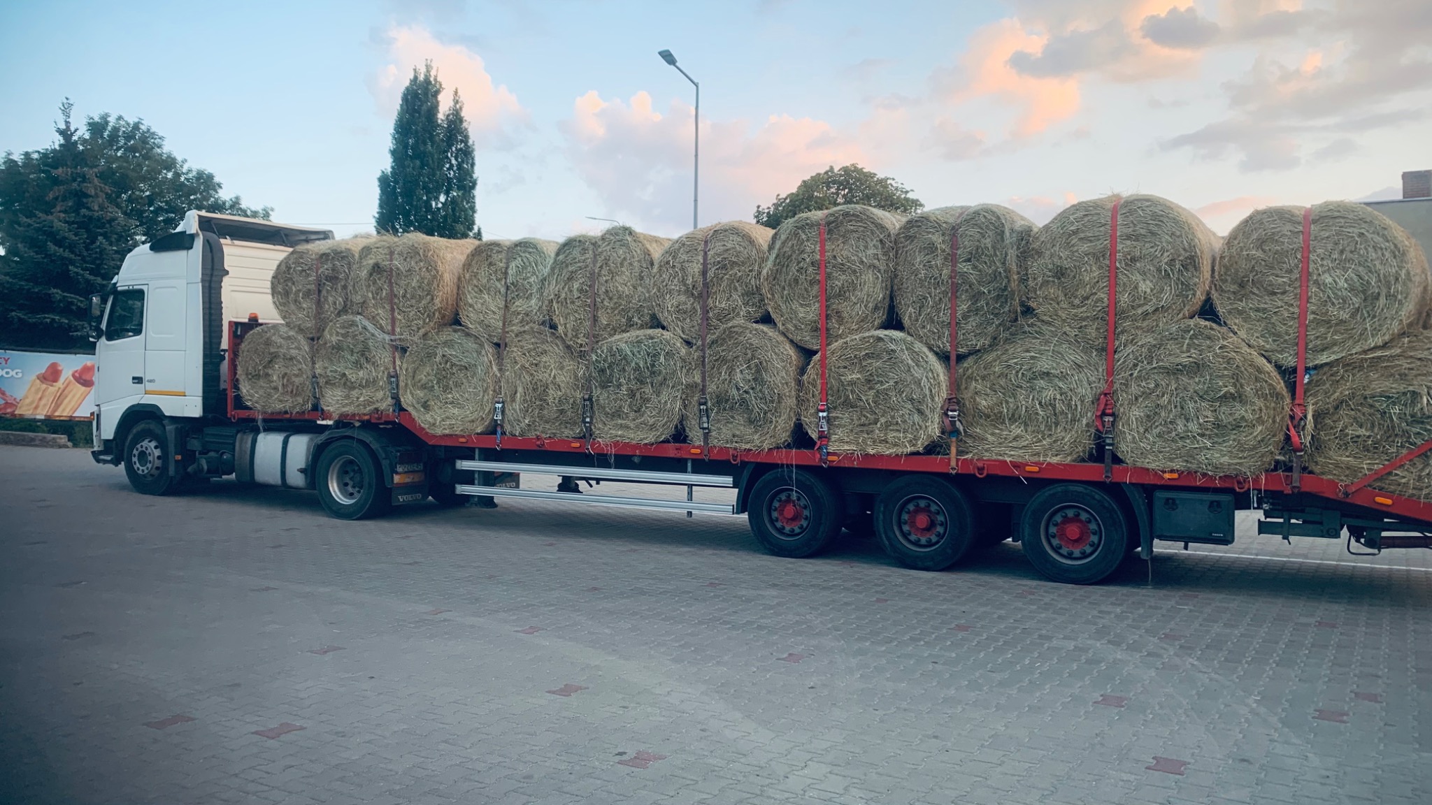Ciężarówka z naczepą załadowana belami siana, zabezpieczonymi czerwonymi pasami transportowymi, zaparkowana na brukowanym placu o zmierzchu.