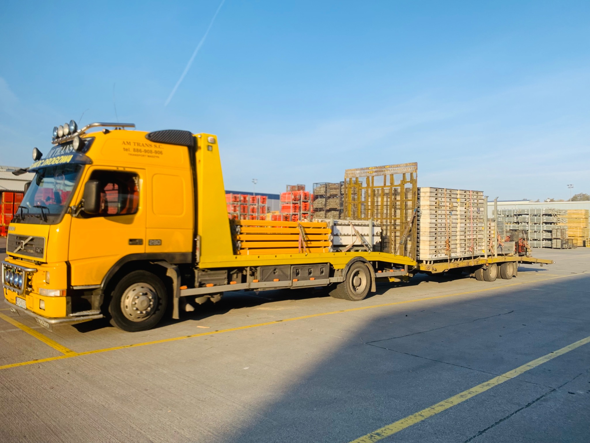Żółta laweta Volvo z ładunkiem metalowych konstrukcji szalunkowych na przyczepie, zaparkowana na betonowym placu w słoneczny dzień.