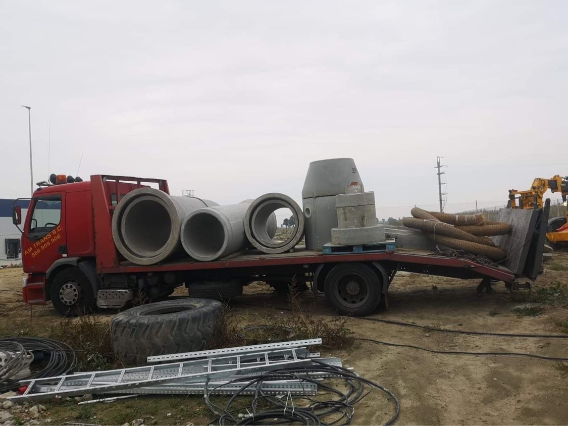 Czerwona ciężarówka z platformą załadowana betonowymi rurami, studzienkami i elementami kanalizacyjnymi na palecie, z widoczną drabiną, oponą i kablami na ziemi.