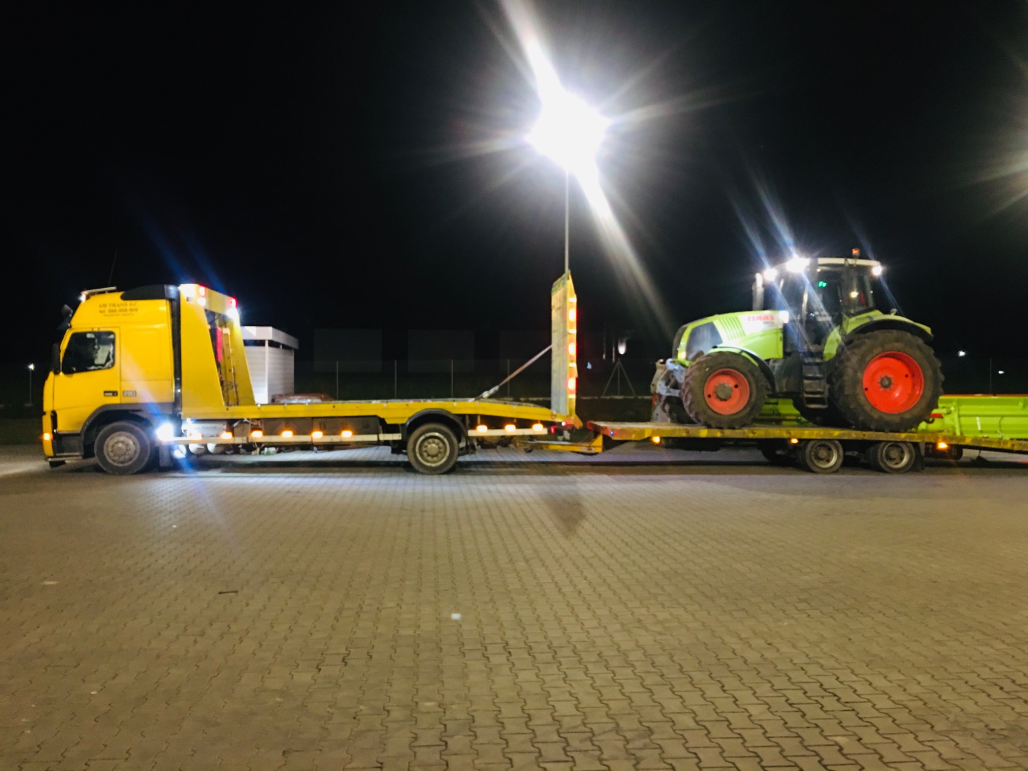 Żółta laweta przewozi zielony traktor Claas na platformie oświetlonej przez lampy uliczne w nocy.