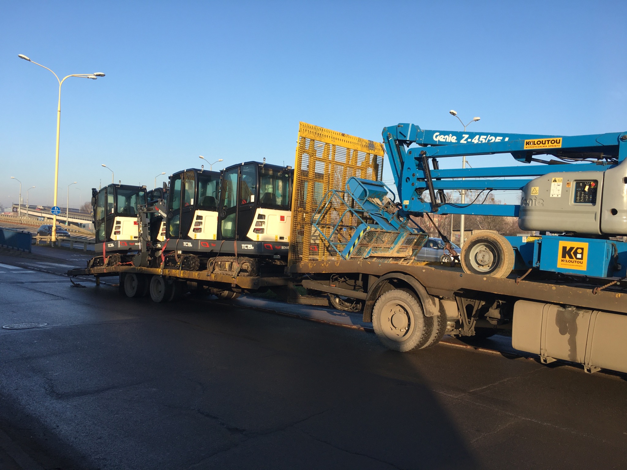 Ciężarówka z platformą przewozi trzy mini koparki Schaeff i podnośnik koszowy Genie Z-45/25 na tle miejskiego krajobrazu z latarniami.