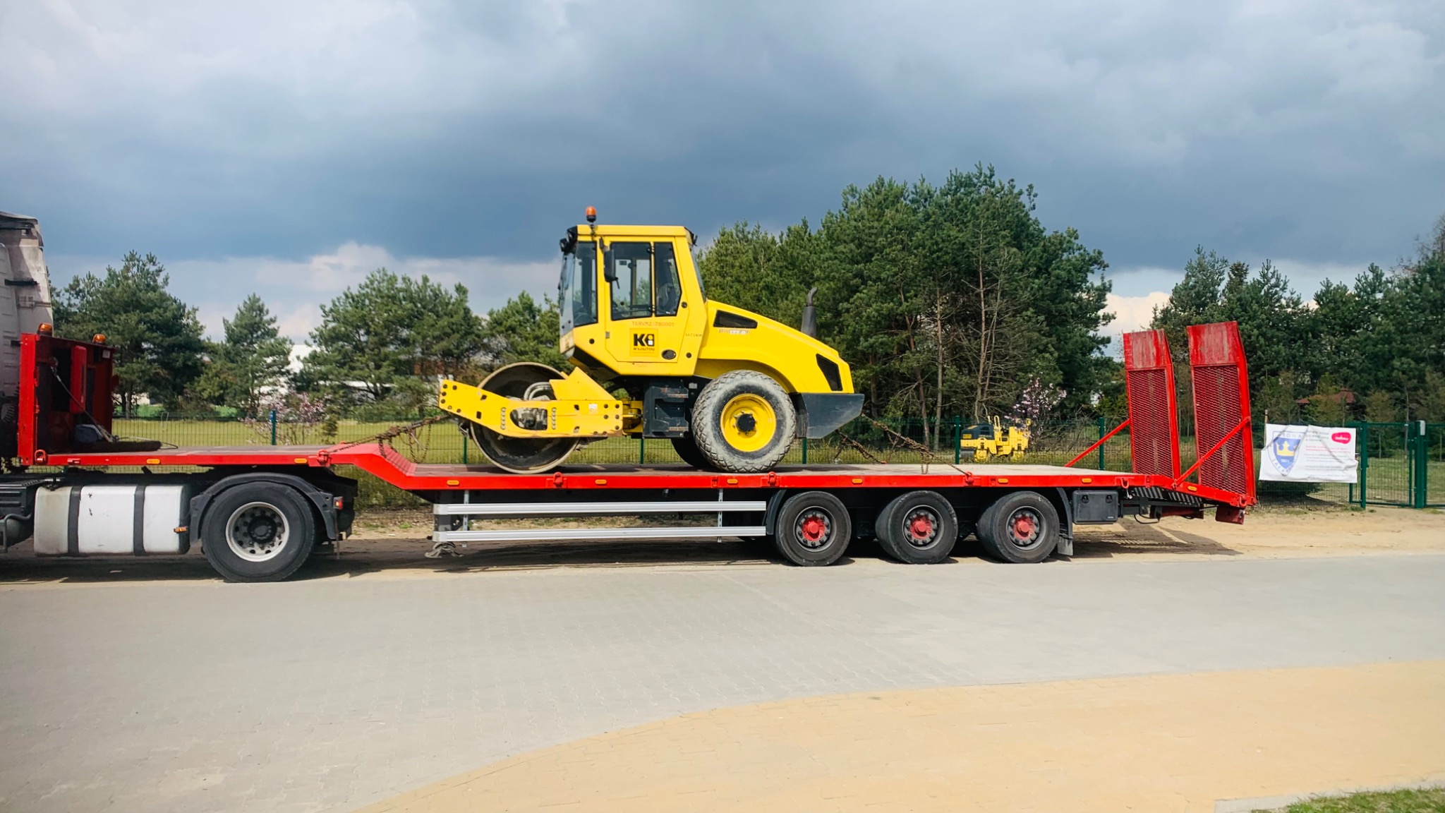 Żółty walec drogowy marki KB przewożony na czerwonej platformie niskopodwoziowej, zabezpieczony łańcuchami, widok z boku na tle zielonych drzew i pochmurnego nieba.