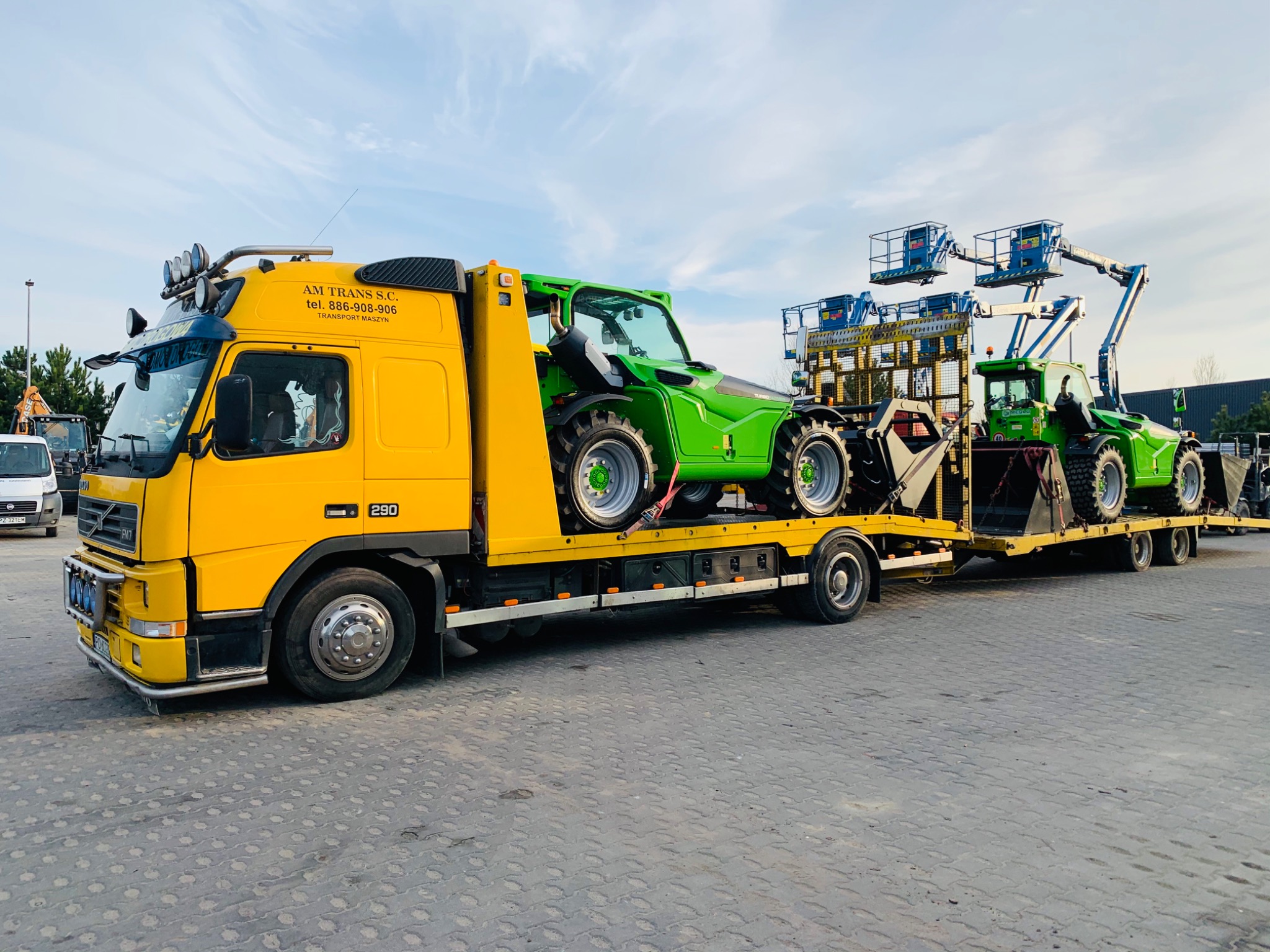 Żółta laweta Volvo przewozi dwa zielone podnośniki koszowe i traktor, widoczne logo firmy AM TRANS S.C. na kabinie, błękitne niebo w tle.