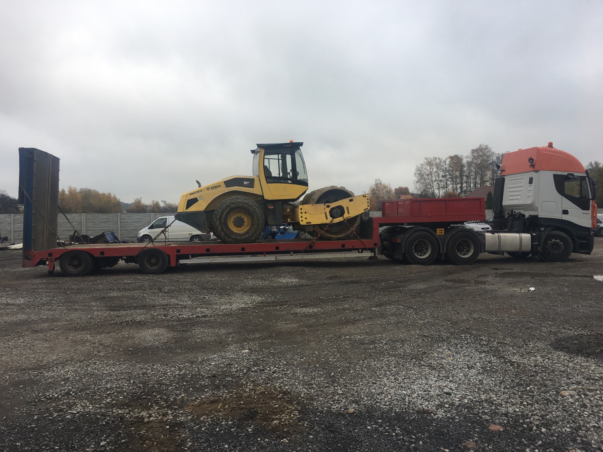 Żółty walec drogowy marki Bomag transportowany na niskopodwoziowej platformie ciągniętej przez ciężarówkę Iveco z pomarańczową kabiną, na szutrowym placu w pochmurny dzień.