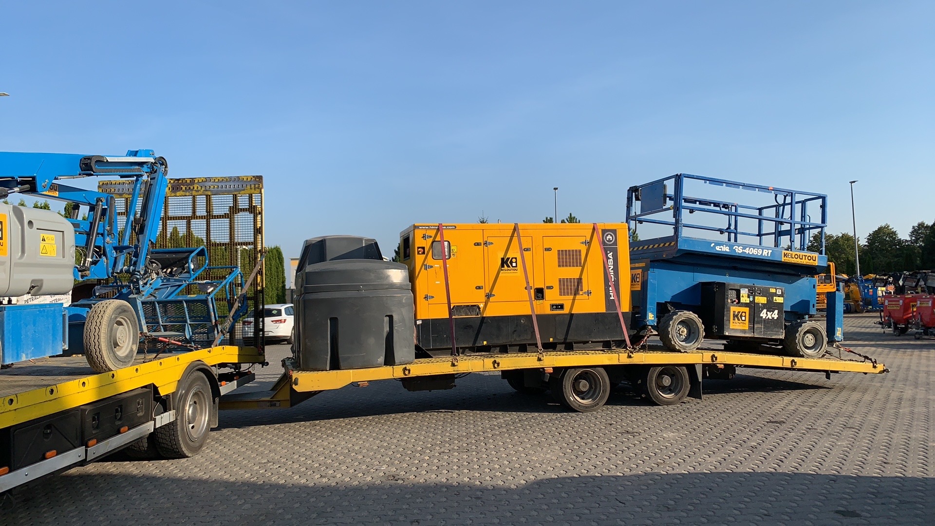 Żółta laweta z dwoma podnośnikami koszowymi i agregatem prądotwórczym, zabezpieczonymi pasami transportowymi.