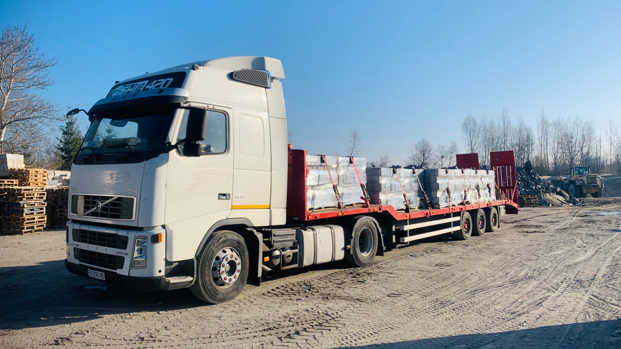 Biała ciężarówka Volvo FH12 z naczepą przewożąca palety z kostką brukową, na placu budowy z widoczną koparką w tle.