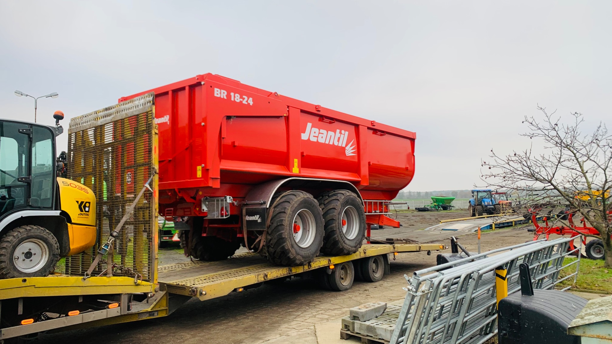 Czerwona przyczepa rolnicza marki Jeantil BR 18-24, z dużymi oponami, umieszczona na żółtej platformie transportowej, zabezpieczona pasami, z widocznym w tle żółto-czarnym wózkiem widłowym i innymi...