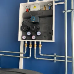 Rozdzielacz ogrzewania podłogowego w białej obudowie z widocznymi pompami obiegowymi, manometrami, zaworami i rurami na tle niebieskiej ściany, z fragmentem zbiornika na dole kadru.