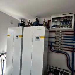 InstAll Technika Grzewcza i Sanitarna - Dwie białe pompy ciepła Rotenso z osprzętem i instalacją hydrauliczną w kolorze niebieskim i czerwonym, zamontowane w pomieszczeniu technicznym. Widoczny rozdzielacz ogrzewania.