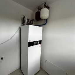 InstAll Technika Grzewcza i Sanitarna - Biała pompa ciepła Viessmann w rogu pomieszczenia z szarymi ścianami i podłogą. Nad nią zbiorniki wyrównawcze i instalacja. Widoczna żarówka na suficie.