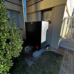 InstAll Technika Grzewcza i Sanitarna - Czarna pompa ciepła Viessmann na betonowych podstawach, z chromowanym kominem, zamontowana przy beżowej ścianie budynku z oknem i zielonym krzewem.