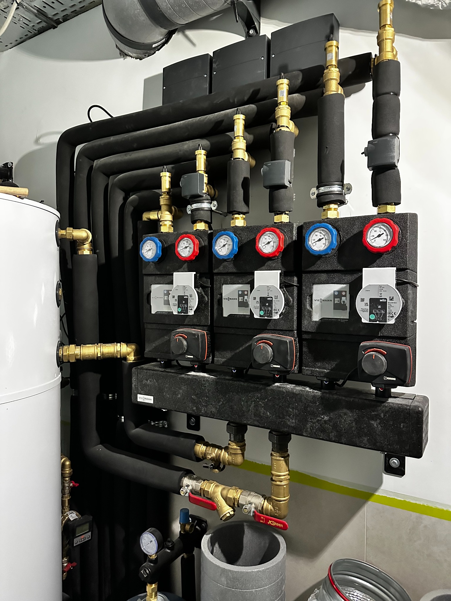 Złożona instalacja grzewcza z czarnymi rurami, manometrami (czerwone i niebieskie) i pompami obiegowymi Wilo, zamontowana na ścianie z szarą listwą przypodłogową.