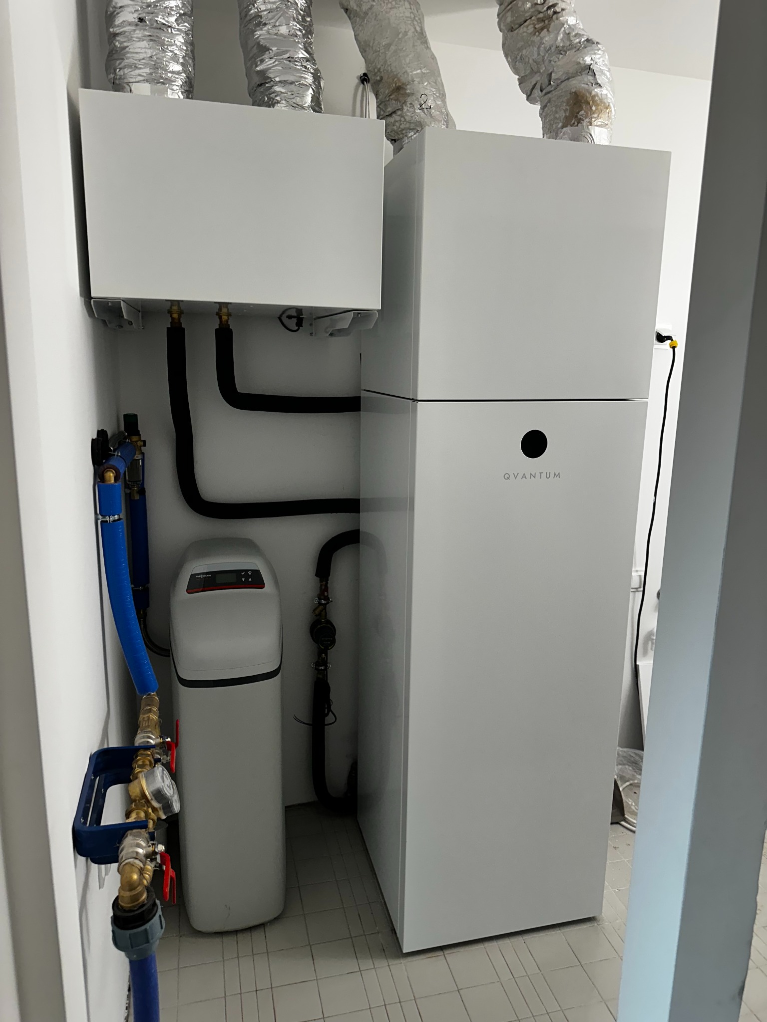 Biała pompa ciepła Quantum z instalacją i filtrem wody w pomieszczeniu. Widoczne rury wentylacyjne i elementy hydrauliczne. Minimalistyczny design, jasne wnętrze.