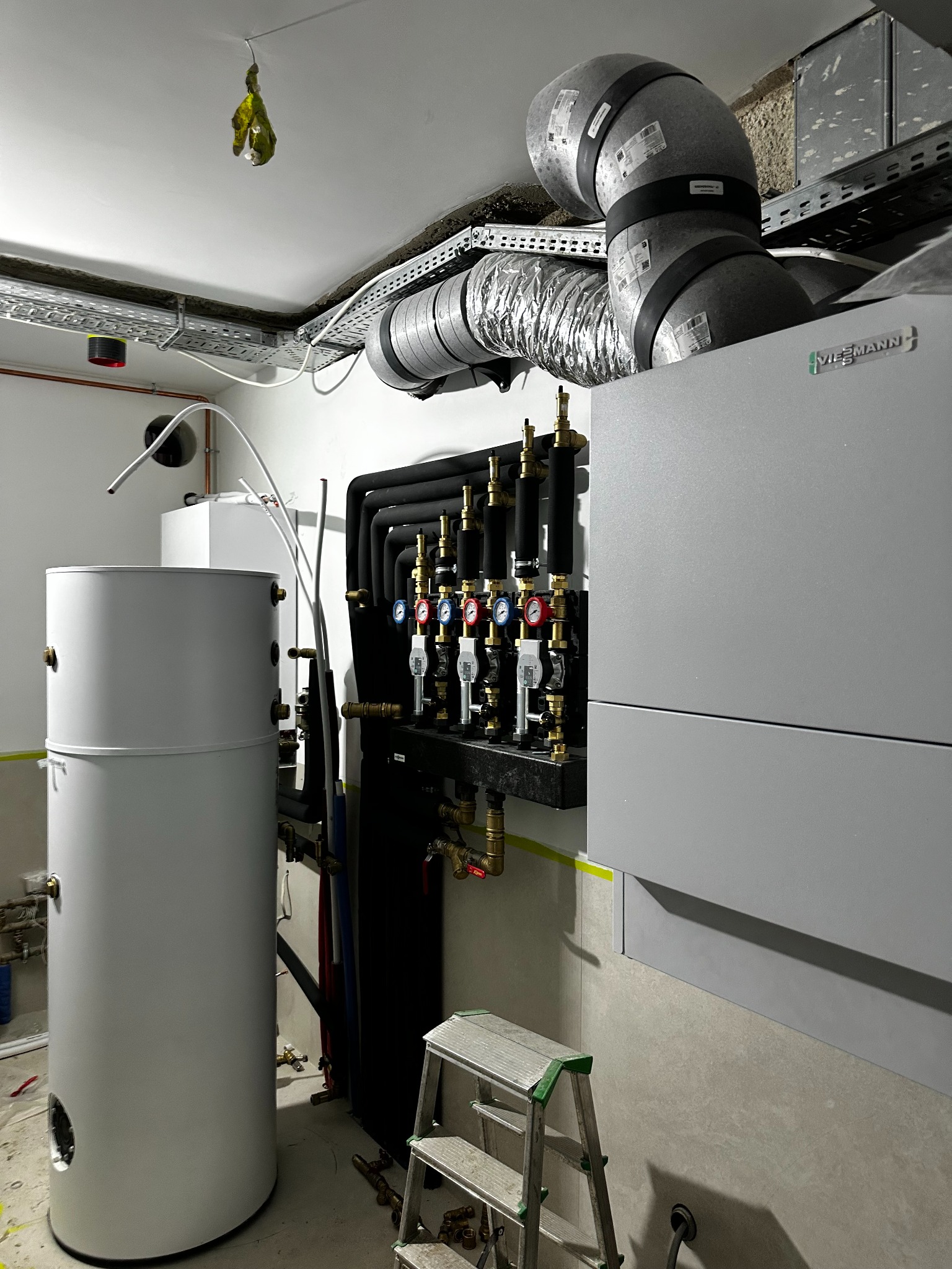Szara kotłownia z zbiornikiem buforowym, rurami, zaworami i piecem Viessmann. Widoczna drabina i instalacje pod sufitem. Ujęcie z dołu, oświetlenie sufitowe.
