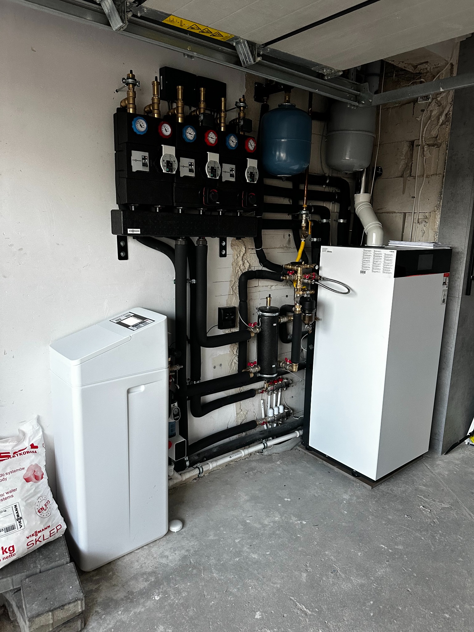 Złożona instalacja grzewcza Viessmann z rozdzielaczem, pompą ciepła i zmiękczaczem wody. Widoczne liczne rury, zawory i manometry. Szara posadzka i surowe ściany.