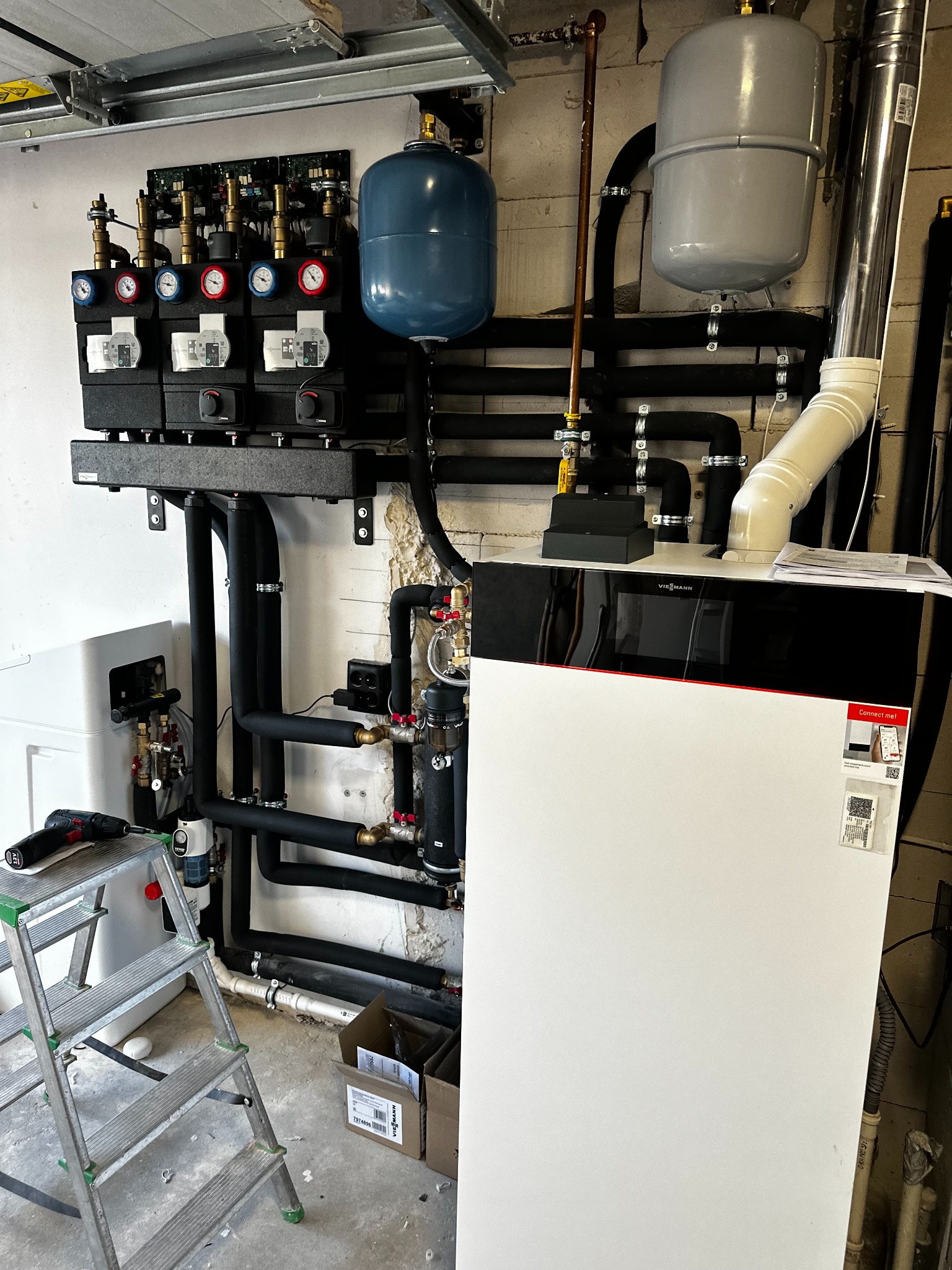 Złożona instalacja grzewcza z piecem Viessmann, rurami w izolacji, zbiornikami wyrównawczymi i rozdzielaczem, widoczna drabina i narzędzia, sugerujące etap montażu.
