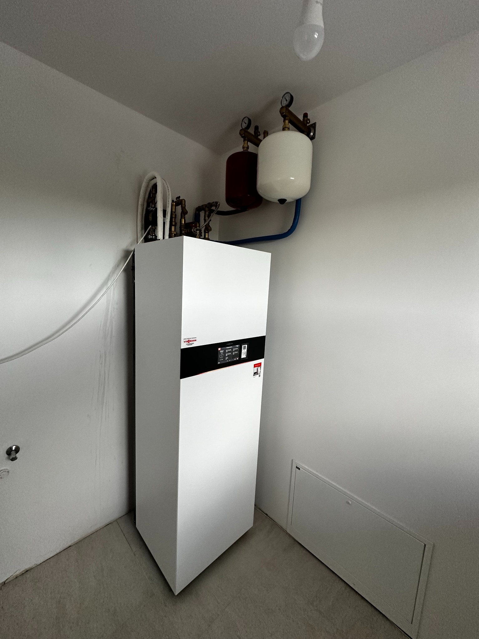 Biała pompa ciepła Viessmann w rogu pomieszczenia z szarymi ścianami i podłogą. Nad nią zbiorniki wyrównawcze i instalacja. Widoczna żarówka na suficie.