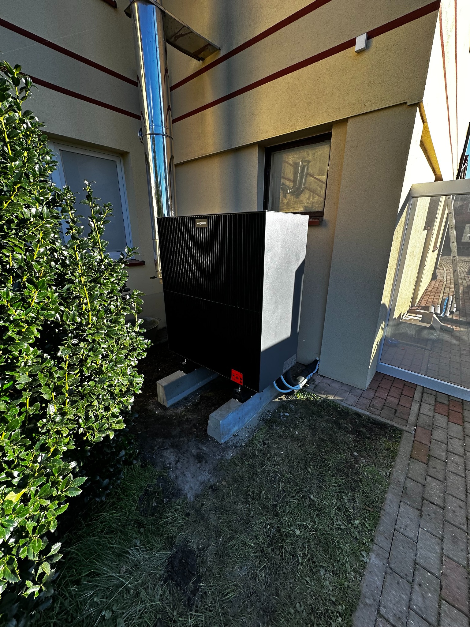 Czarna pompa ciepła Viessmann na betonowych podstawach, z chromowanym kominem, zamontowana przy beżowej ścianie budynku z oknem i zielonym krzewem.
