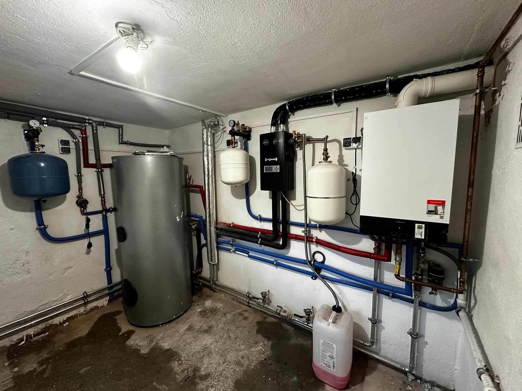 Złożona instalacja grzewcza w kotłowni: zbiornik, piec, naczynia przeponowe, rury miedziane i plastikowe. Widoczny kanister z płynem. Oświetlenie sufitowe.