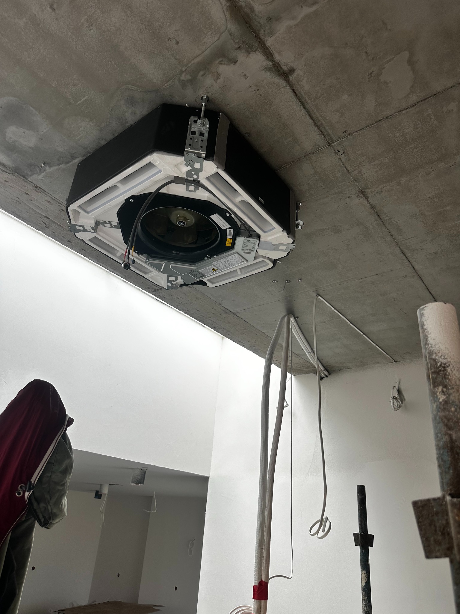 Czarna, kasetonowa jednostka klimatyzacji zamontowana na surowym, betonowym suficie. Widoczne przewody i elementy montażowe. W tle białe ściany w trakcie wykończenia.