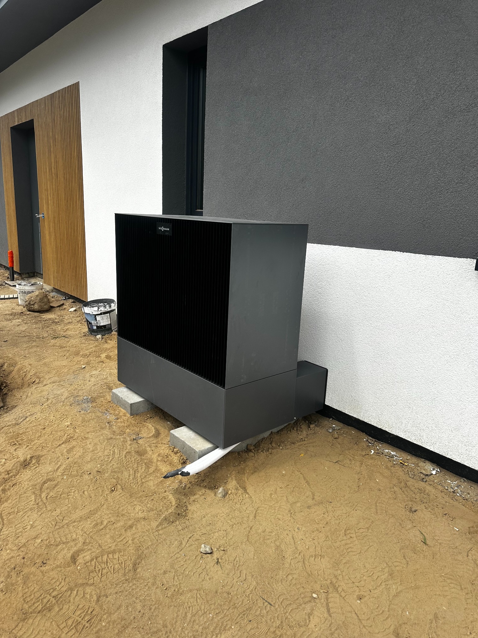 Szara pompa ciepła Viessmann zamontowana na betonowych bloczkach przy elewacji budynku z drewnianymi akcentami. Widoczne rury instalacyjne i teren budowy.