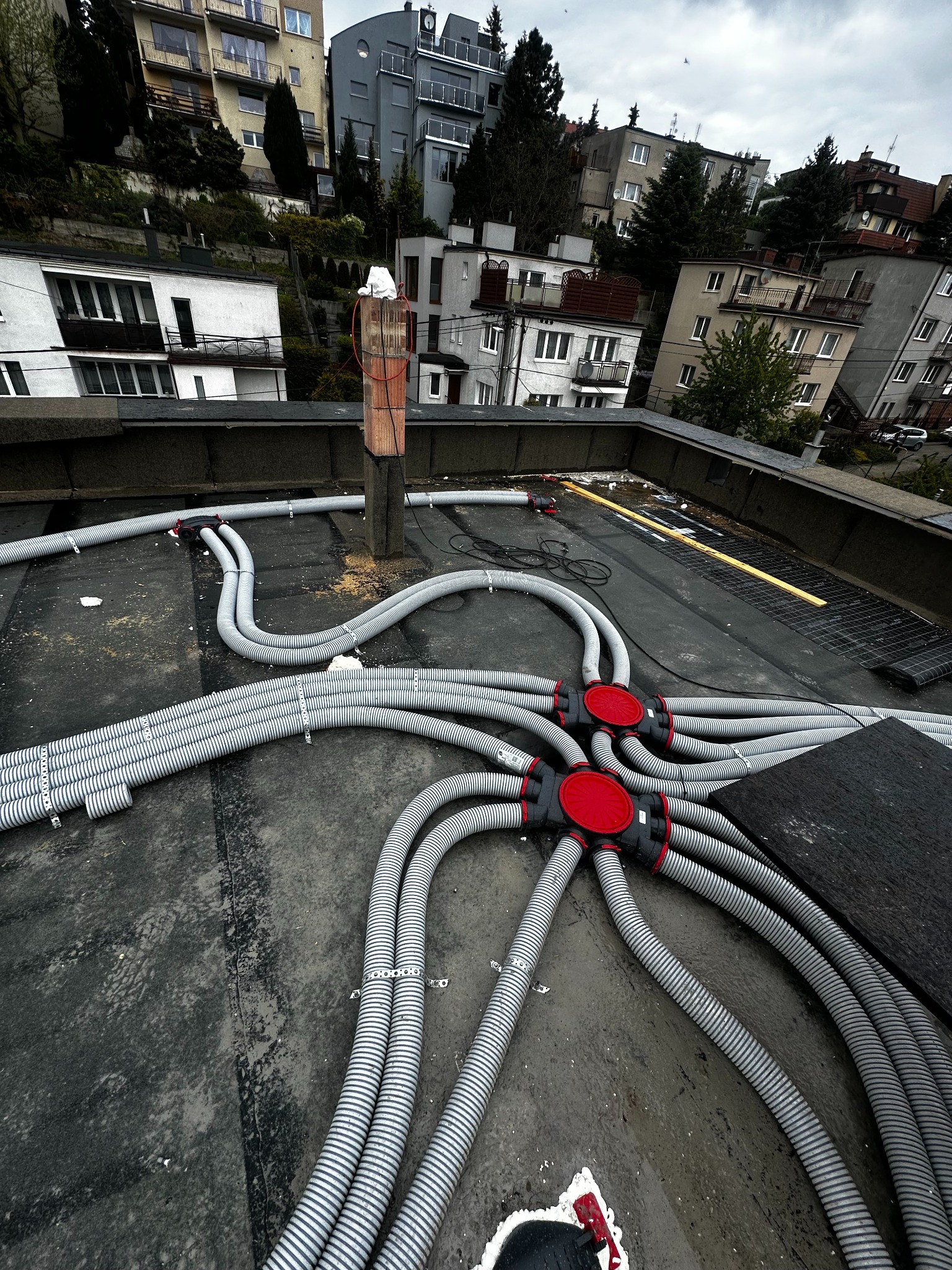 Szare, karbowane rury wentylacyjne z czerwonymi łącznikami na czarnym dachu budynku. Widok z góry, instalacja w trakcie montażu, budynki w tle.