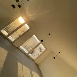 Wnętrze pokoju z trzema oknami, surowy stan wykończenia, widoczne lampy wiszące i sufit z instalacją elektryczną, z perspektywy od dołu.