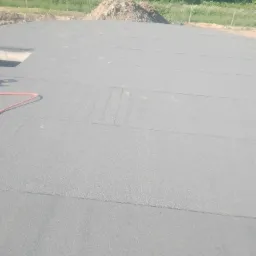 Szara, płaska powierzchnia pokryta papą termozgrzewalną w trakcie prac budowlanych, widoczny fragment czerwonego węża i wykop pod fundamenty.