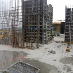 Zbrojenie i szalunki przygotowane do wylewania betonu na budowie, widoczne pionowe pręty zbrojeniowe i metalowe formy szalunkowe, podłoże z betonu z kałużami wody.