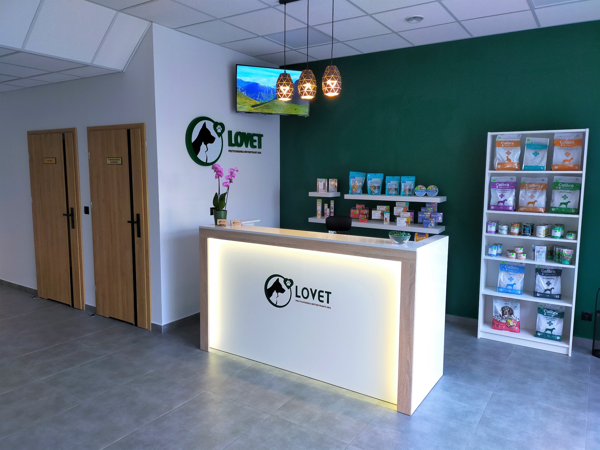 Nowoczesna recepcja w przychodni weterynaryjnej z podświetlanym logo firmy Lovet, ekspozycją karm Calibra na półce i telewizorem wyświetlającym krajobraz górski.