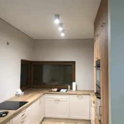 Nowoczesna kuchnia w jasnych barwach z drewnianym blatem, oświetleniem punktowym i zamontowaną płytą indukcyjną. Na blacie widoczne akcesoria kuchenne i środki czystości.