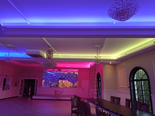 Sala bankietowa z oświetleniem LED w kolorach niebieskim, żółtym i różowym, widoczne żyrandole i drabina oparta o ścianę