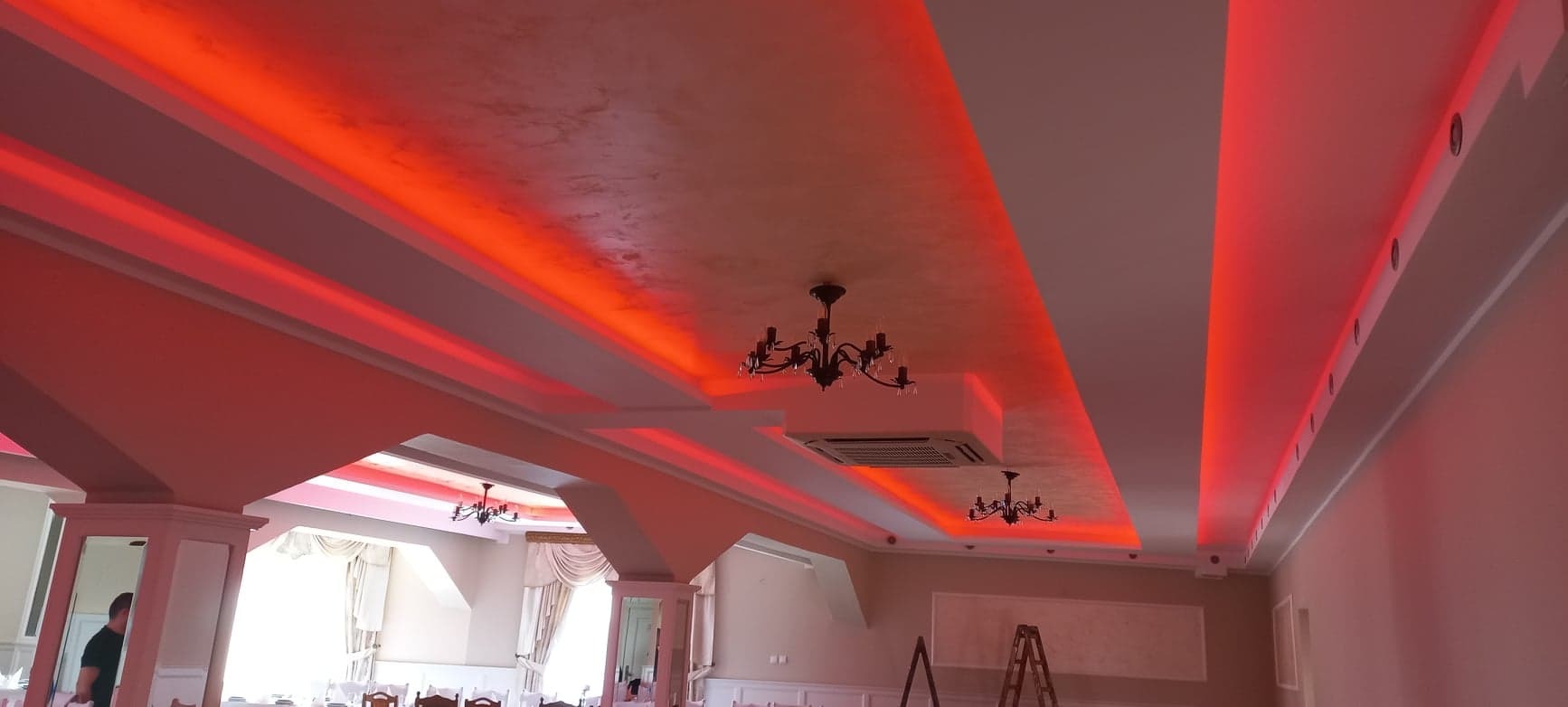 Sufit w sali bankietowej z podwieszanymi kasetonami oświetlonymi czerwonymi LED-ami, widoczne żyrandole i drabina aluminiowa w tle.