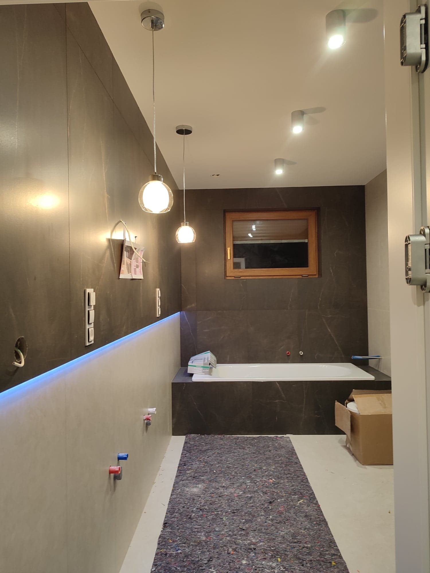 Nowoczesna łazienka w trakcie wykańczania: widoczne podłączenia elektryczne, oświetlenie sufitowe punktowe i wiszące, okno w drewnianej ramie, wanna obudowana płytkami imitującymi marmur...