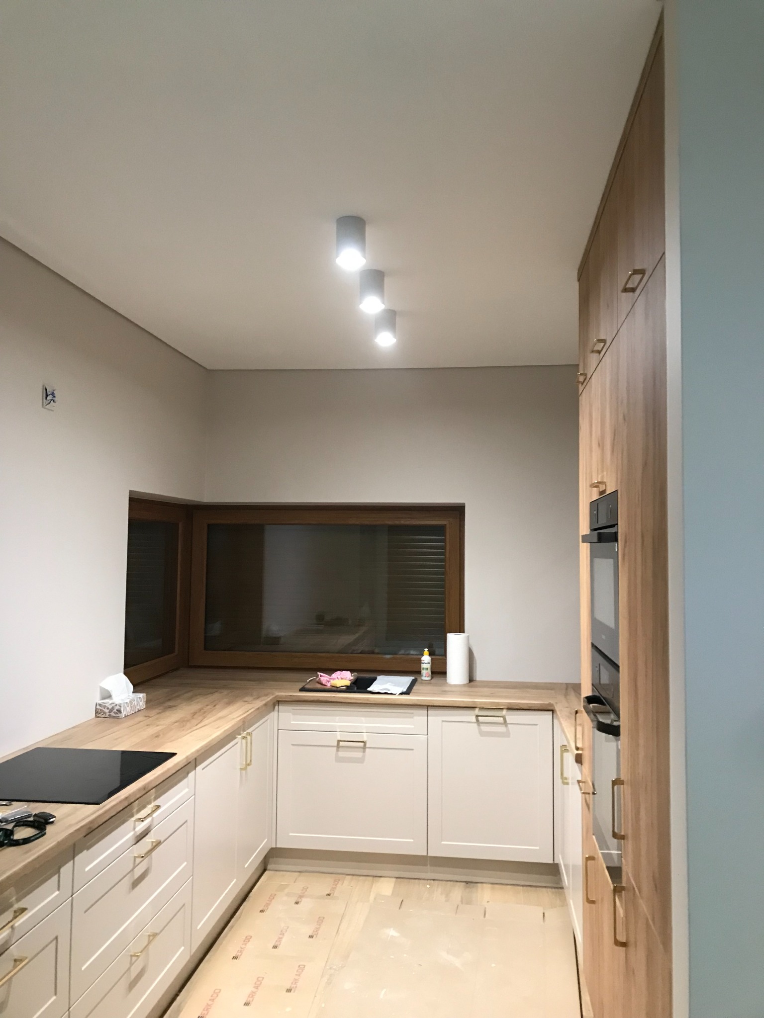 Nowoczesna kuchnia w jasnych barwach z drewnianym blatem, oświetleniem punktowym i zamontowaną płytą indukcyjną. Na blacie widoczne akcesoria kuchenne i środki czystości.