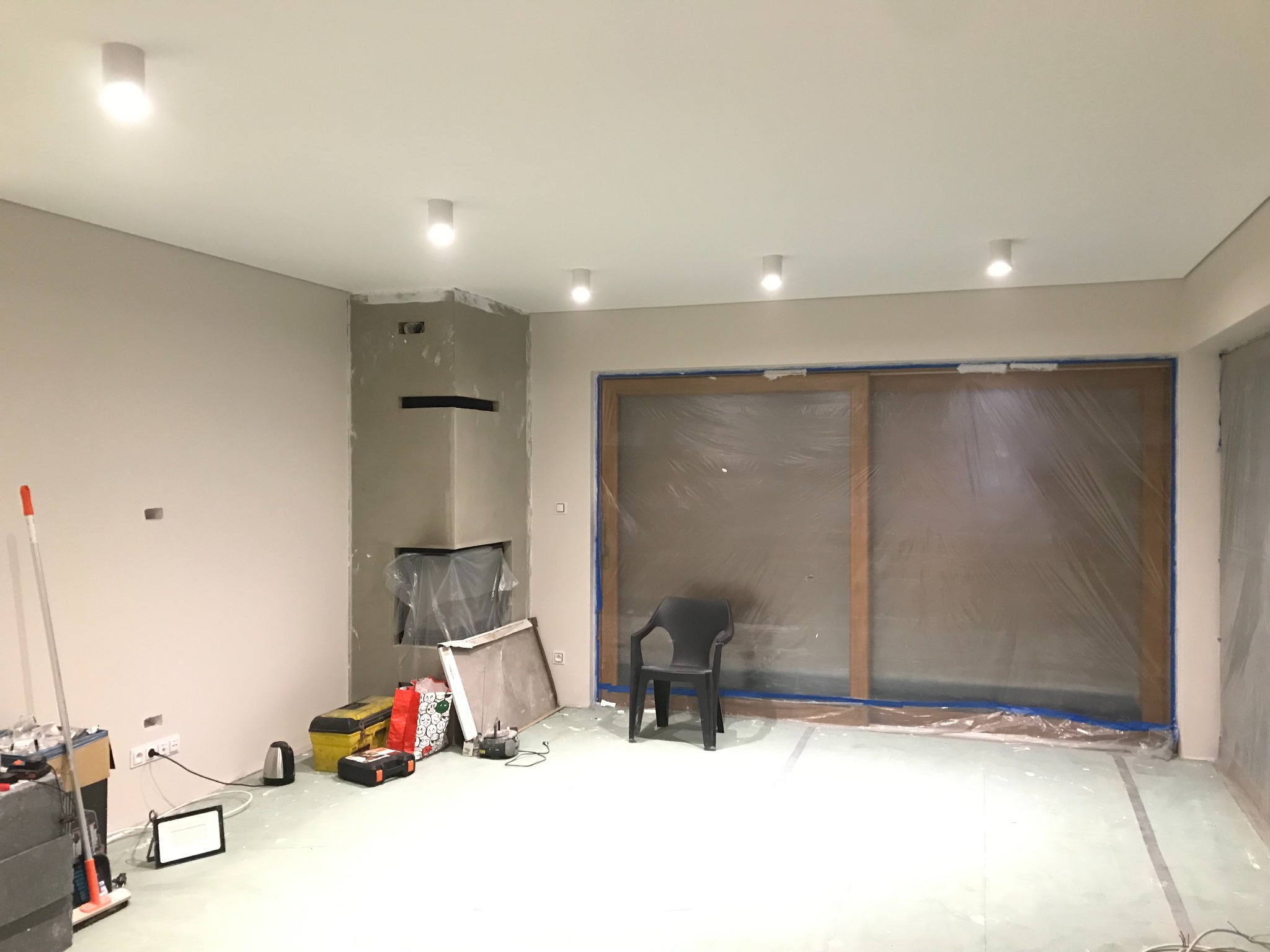 Wnętrze w trakcie prac wykończeniowych: widoczne zamaskowane folią okno przesuwne, surowy kominek, narzędzia i przedłużacze przy ścianie, plastikowe krzesło na środku pomieszczenia,...