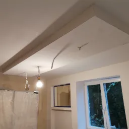 Wykończony sufit podwieszany z płyt kartonowo-gipsowych z widocznymi przewodami oświetleniowymi i lampą wiszącą, okno z widokiem na zieleń za nim, ściana przygotowana do malowania, zakryte folią...